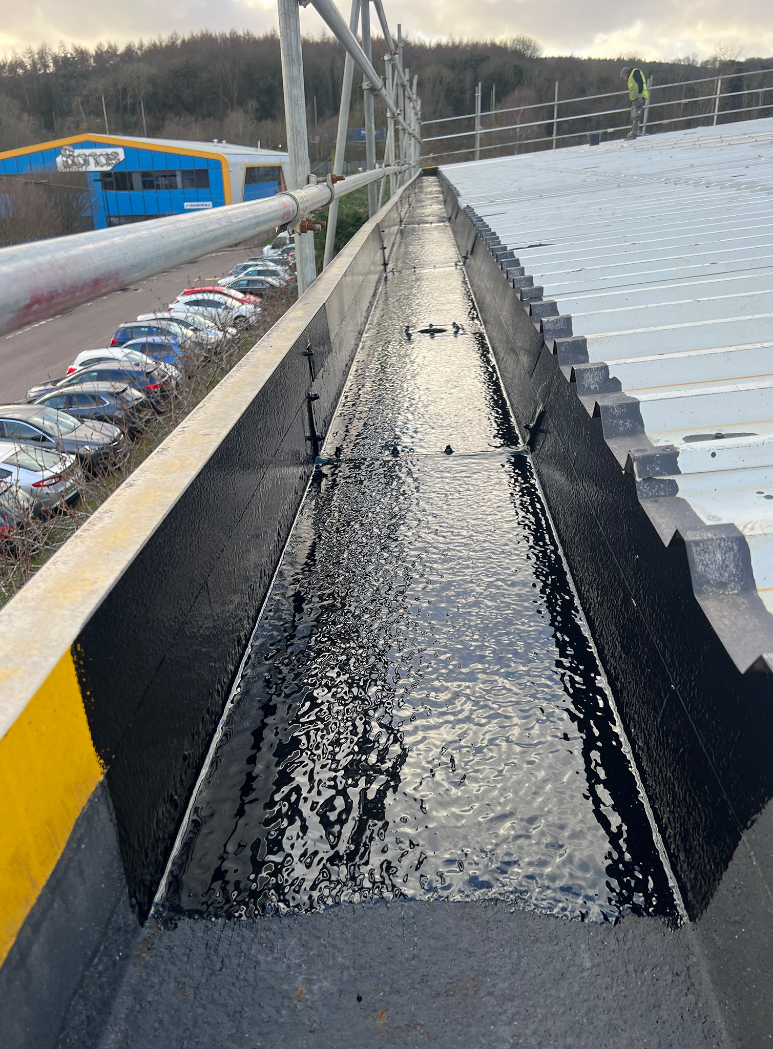 DriSilane ReTrim protective coating being brush-applied in gloss black to the internal and external faces of a steel box gutter, with the wet coat visible across the gutter channel and upper flanges